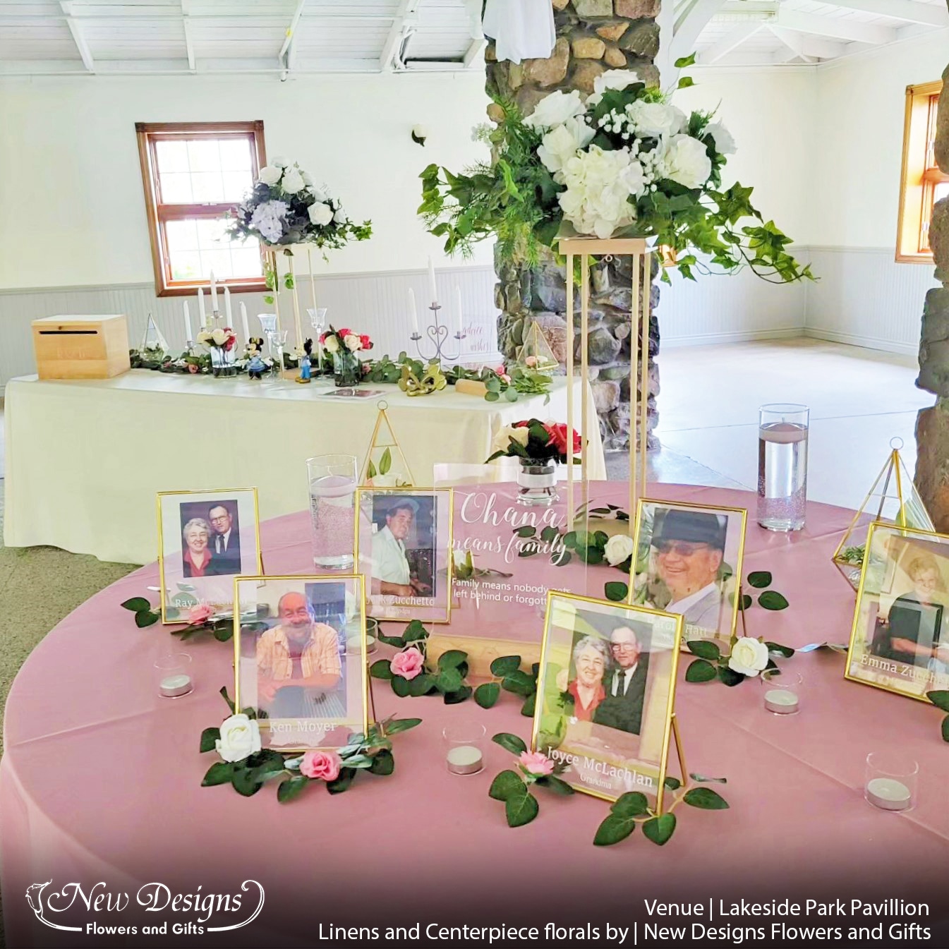 A memorial table and sign in table in a cobblestone pavillion, with dusty pink tablecloths, and a tall centerpiece on a 4 posted wooden frame, topped with white flowers and greenery